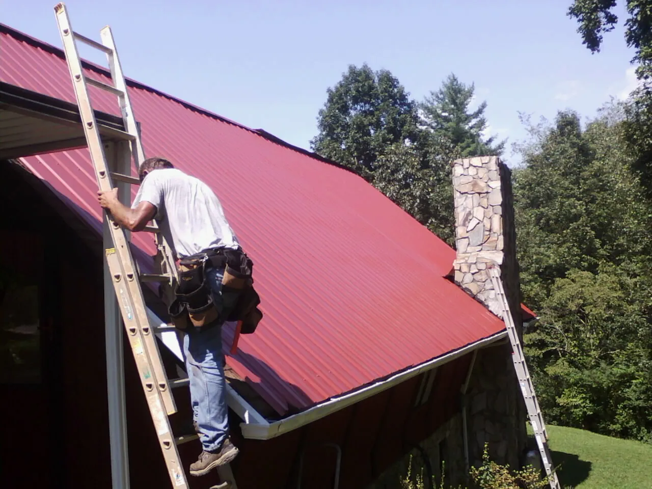 Roofer on a ladder beside a red metal roof and stone chimney — Soffit & Fascia Repair by Roofing Brothers in Yarmouth Port